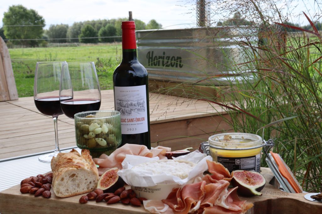 apéritif sur la terrasse de la tiny house avec vin bordelais et une planche de fromages & charcuterie. On observe les verres de vin remplis et le bain nordique en arrière plan